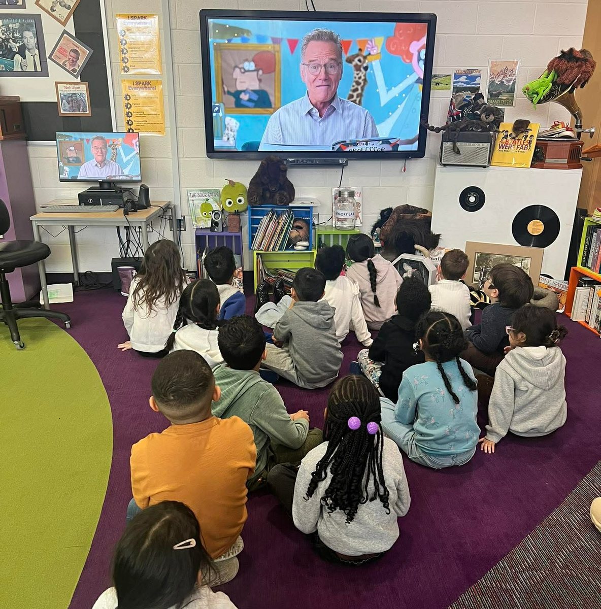 A class sitting on the floor watching Bryan Cranston read Professor Nincompoop on the classroom screen